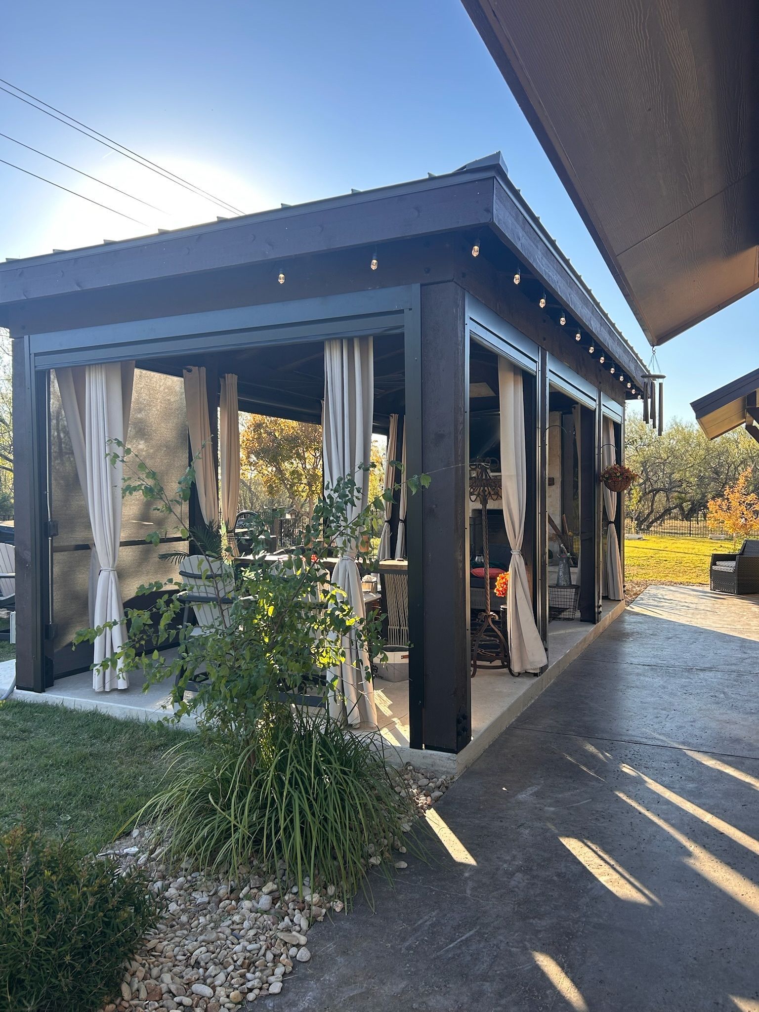 Outdoor gazebo with dark wood frame, light curtains, and a concrete patio.