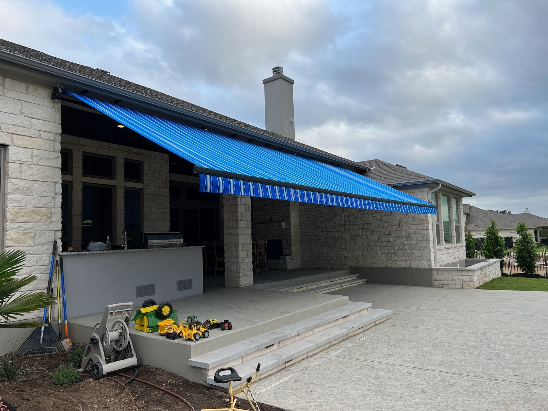Blue and white striped awning over a stone patio, with tools and a house in the background.