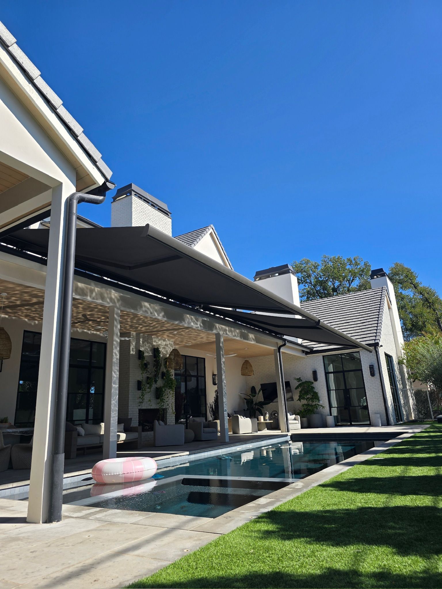 Backyard with a pool and covered patio. White house, green grass, and a bright blue sky.
