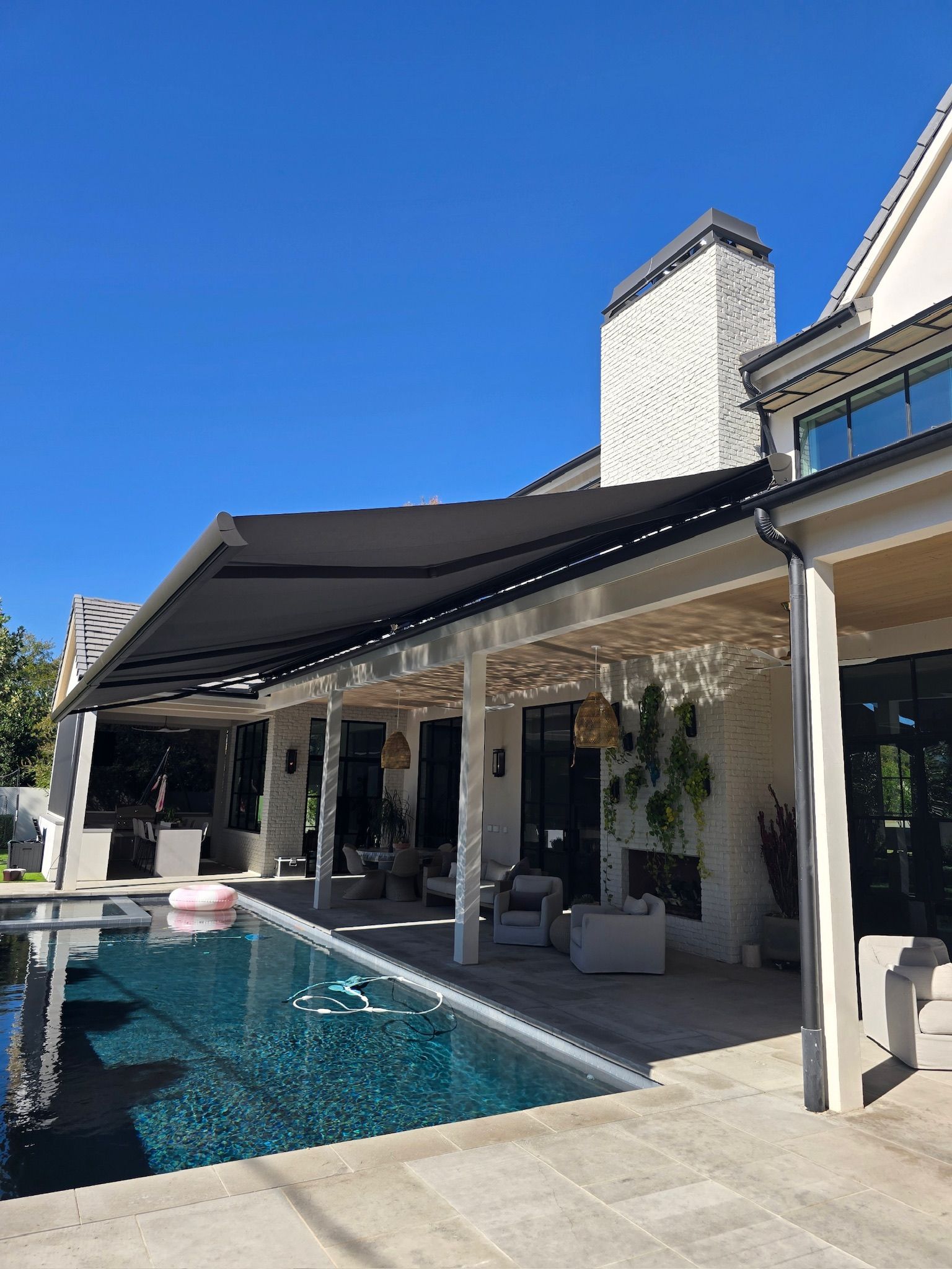 Poolside patio with retractable dark awning, near a large stone house on a sunny day.