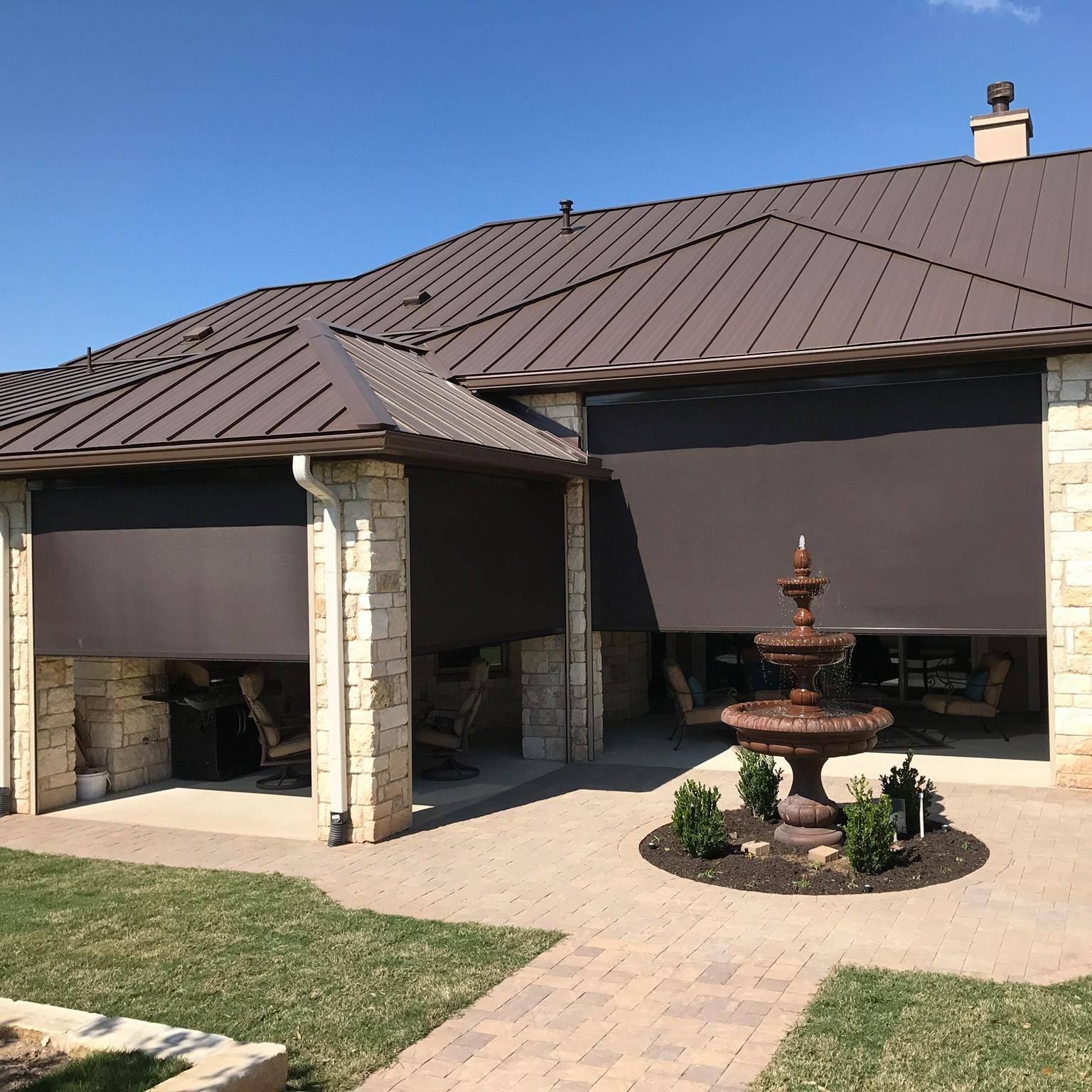 Exterior view of a house with brown metal roof, stone pillars, and retractable sunshades, featuring a fountain on the patio.