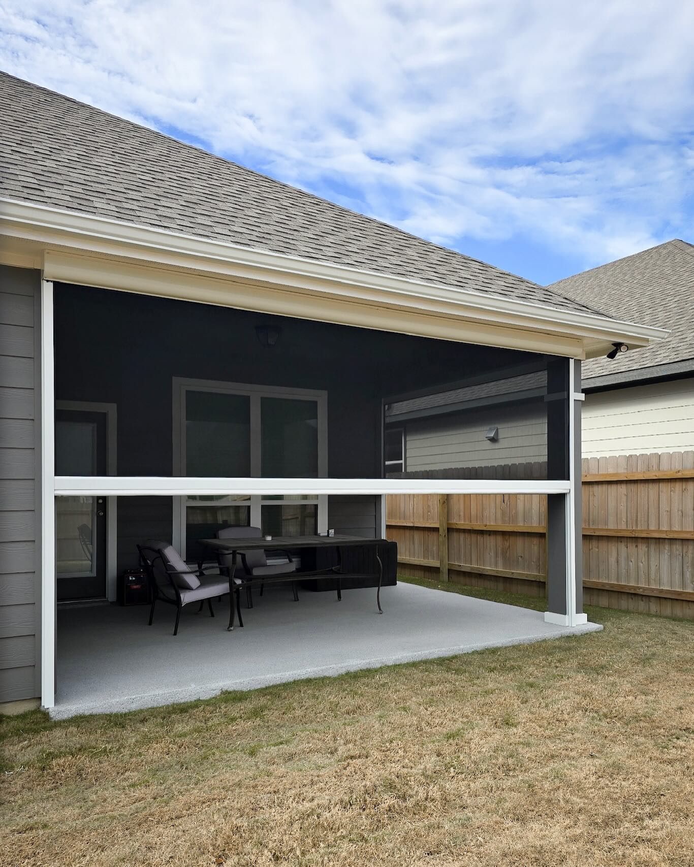 Covered patio with retractable screen. Outdoor seating on concrete patio, enclosed by a gray house and fence.