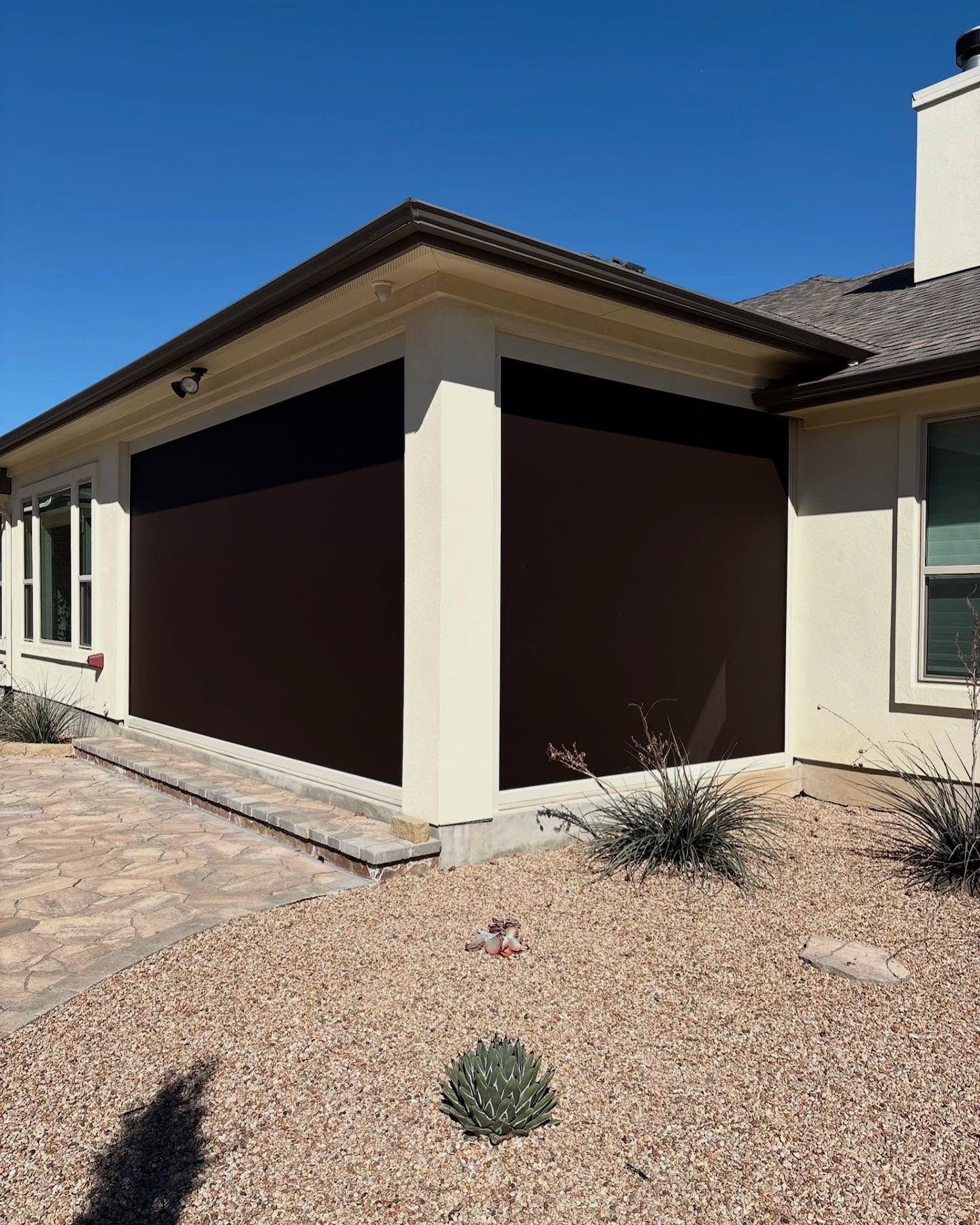 Exterior view of a house with brown sun shades deployed on an outdoor patio. The sky is clear blue.