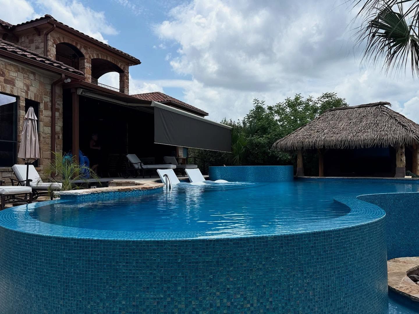 Pool with blue tile and a thatched roof bar under a cloudy sky.