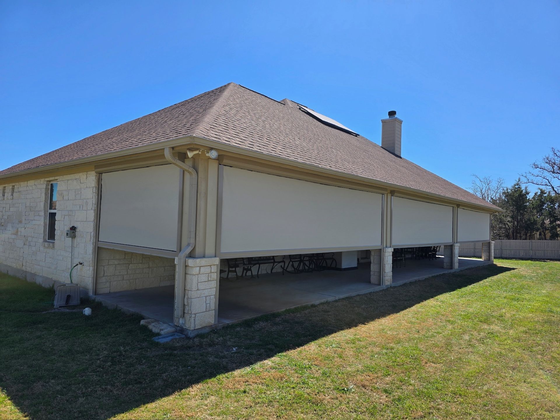 Tan house with lowered beige sun shades on a patio, under a brown shingle roof, on a sunny day.