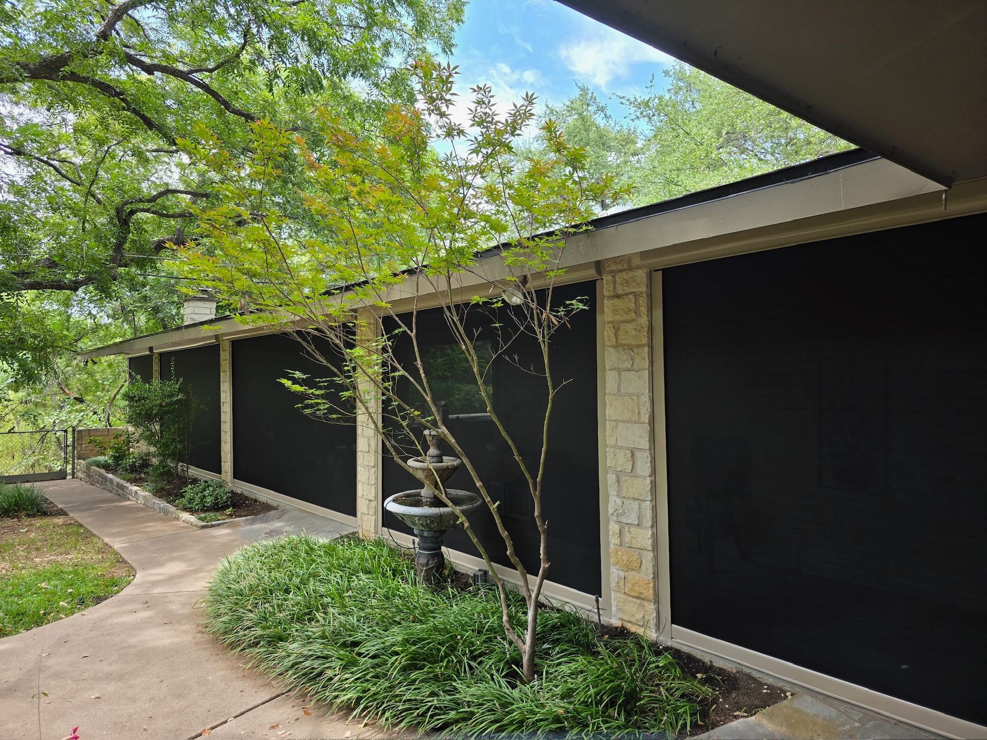 Long exterior wall with black screens, stone columns, plants, and a walkway.