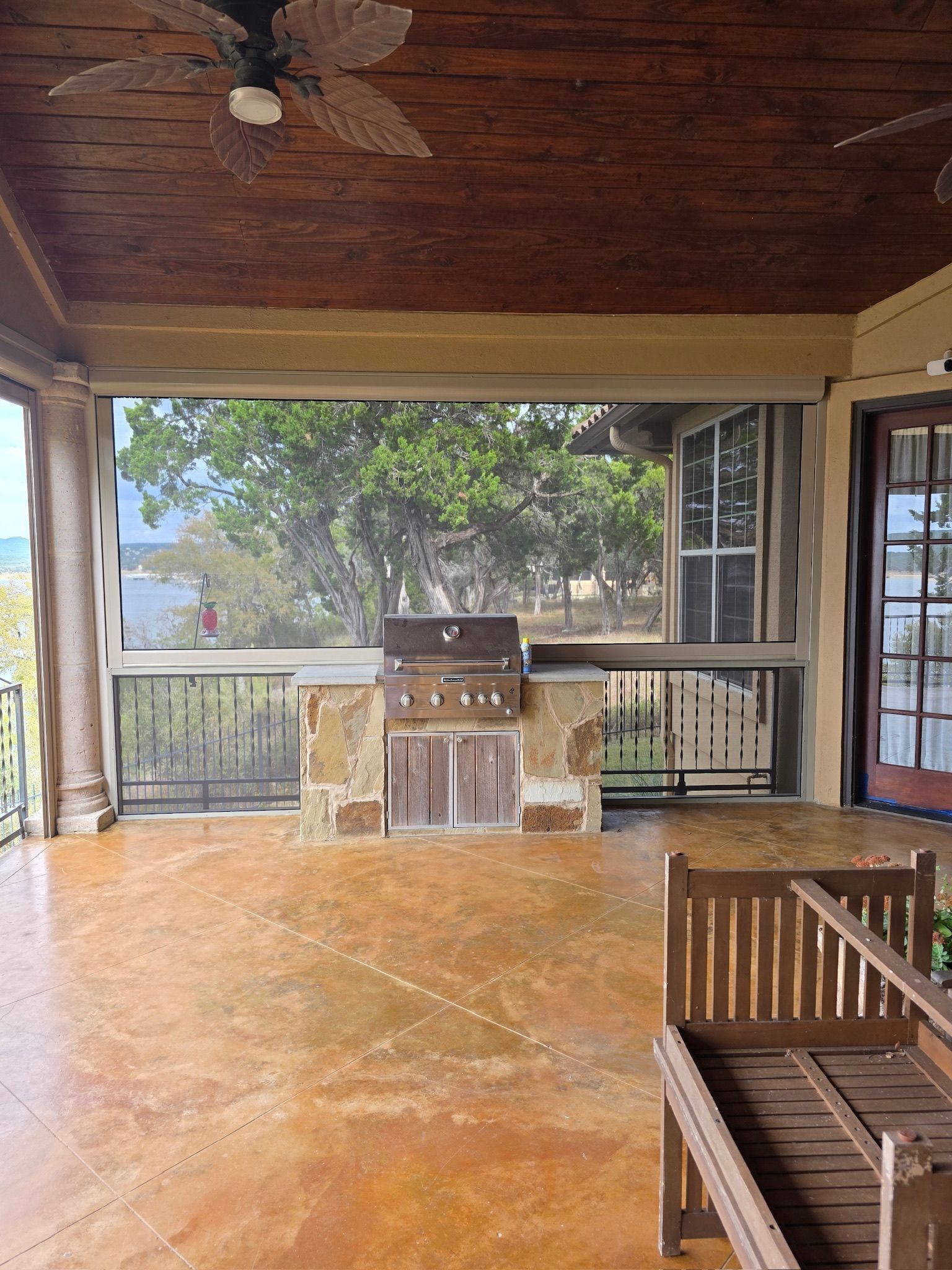 Patio with a built-in grill, retractable screen, and wooden furniture, overlooking a lake.
