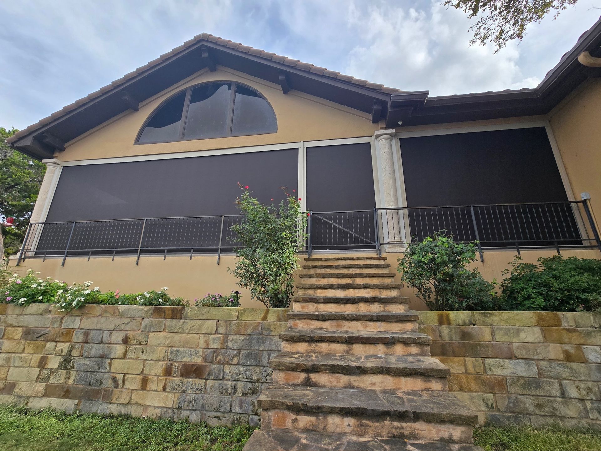 Beige house with arched window and black outdoor shades; stone steps lead up to a patio.
