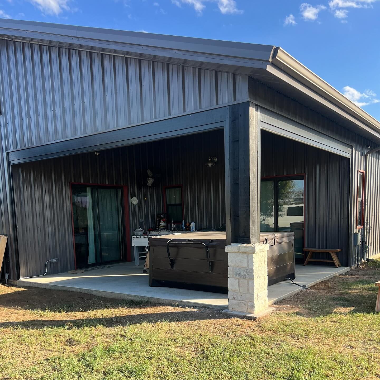 A metal-sided building with a covered patio. A hot tub sits under the overhang. Blue sky is visible.