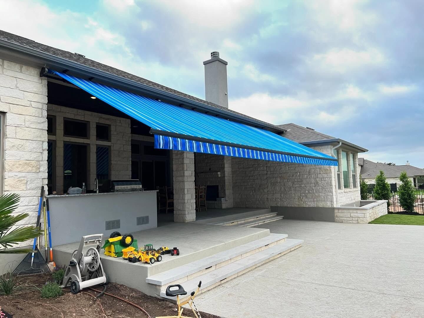 Blue striped retractable awning over a concrete patio outside a light stone house.