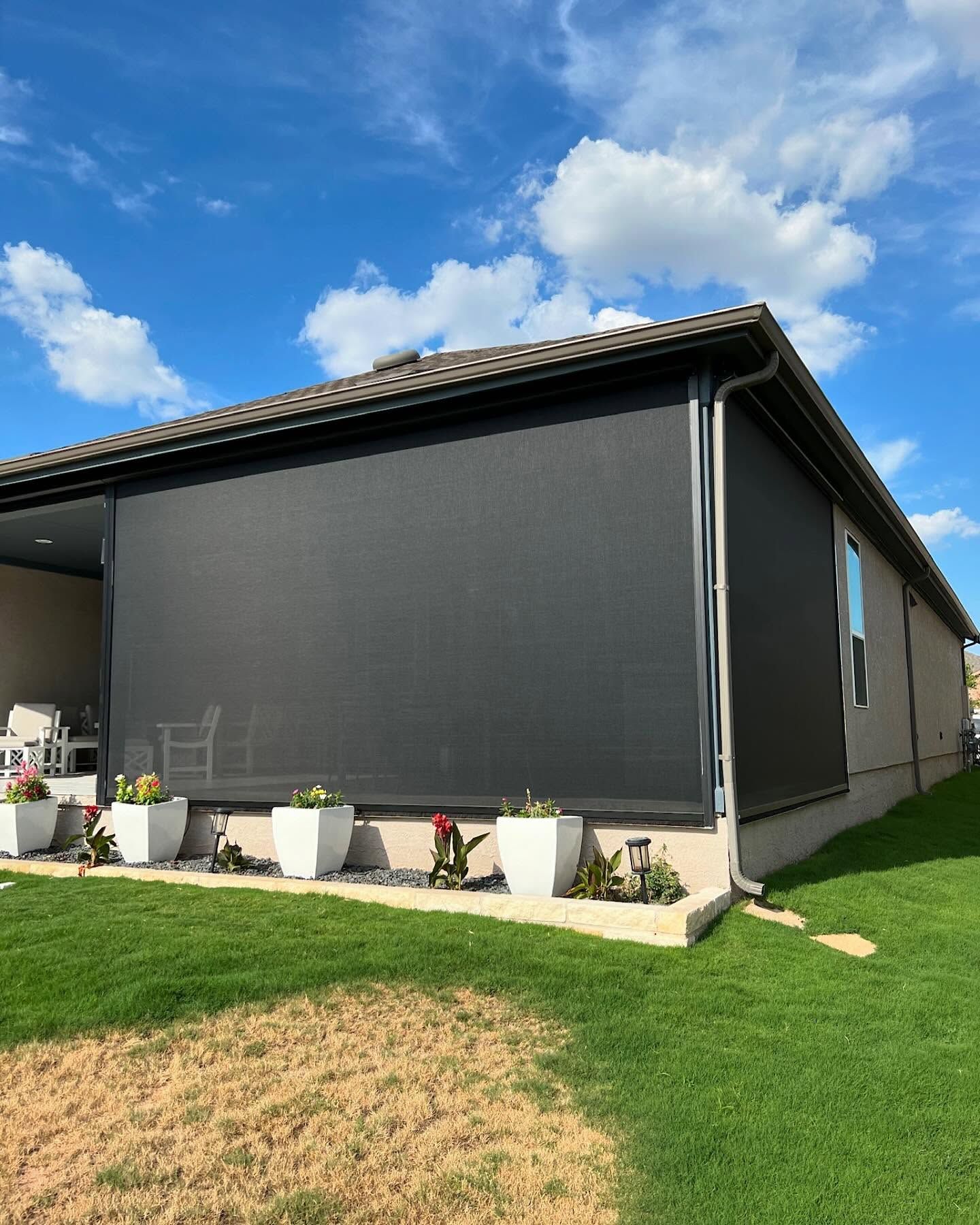 Black sun shades on a house, flowers in white planters line the patio edge. Blue sky with clouds.