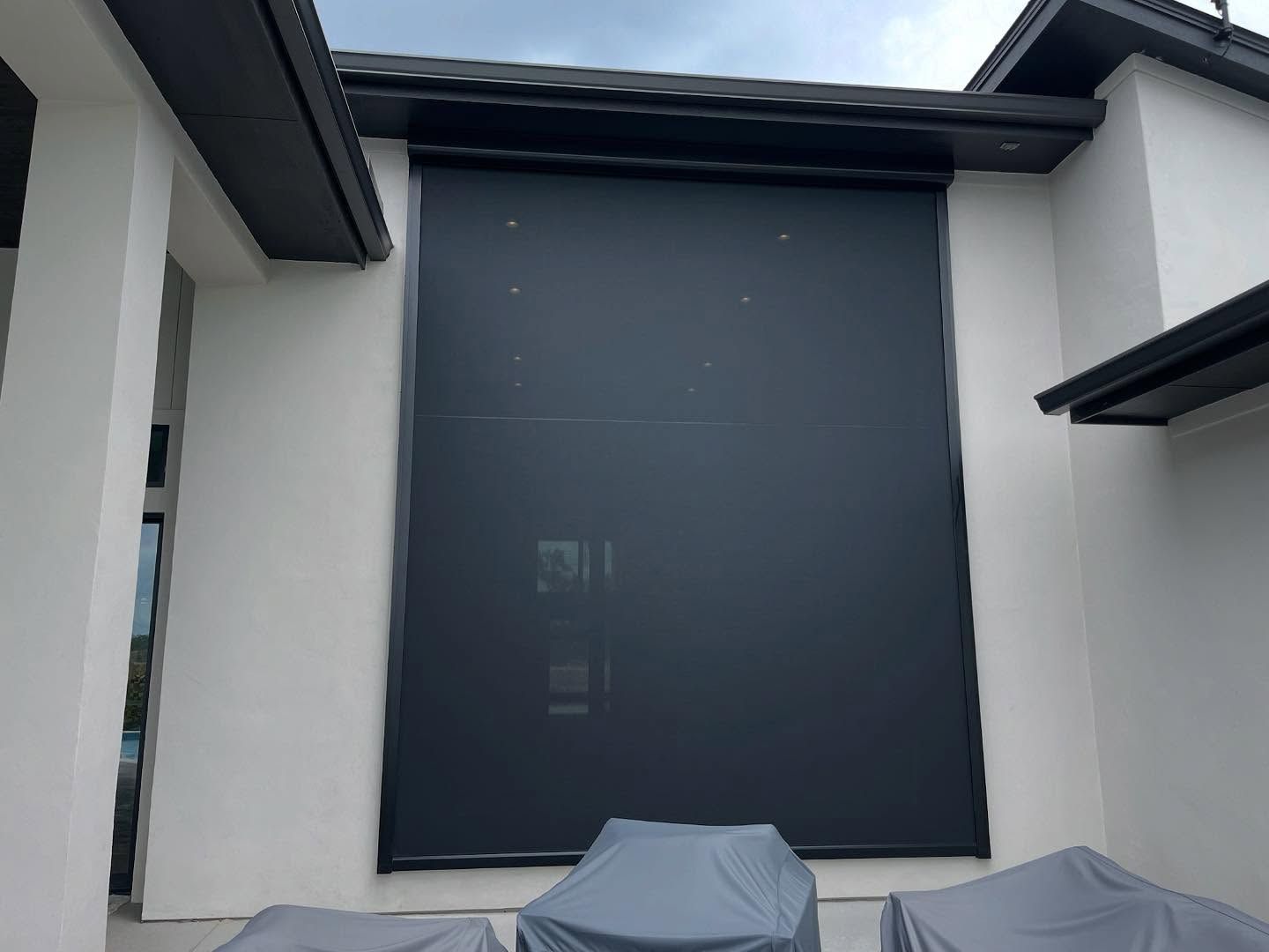 Black outdoor roller shade on a white building, covering patio area with covered furniture.