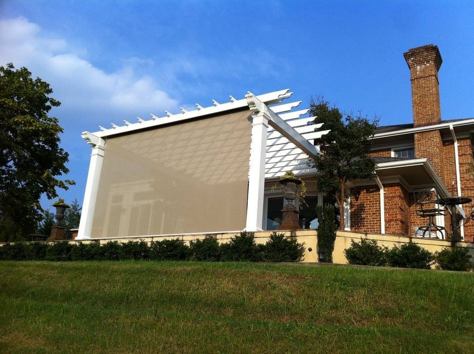 Beige outdoor shade attached to a white pergola on a brick house; sunny day.