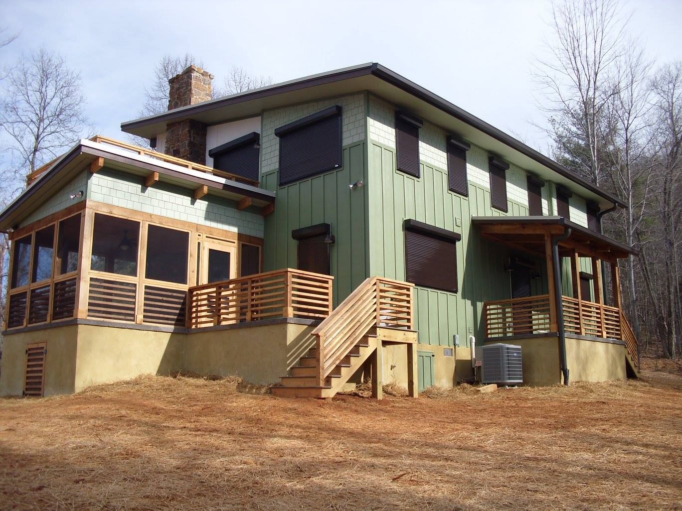 Two-story house with green siding, wooden decks, stairs, and dark window shutters. Set in a wooded area.