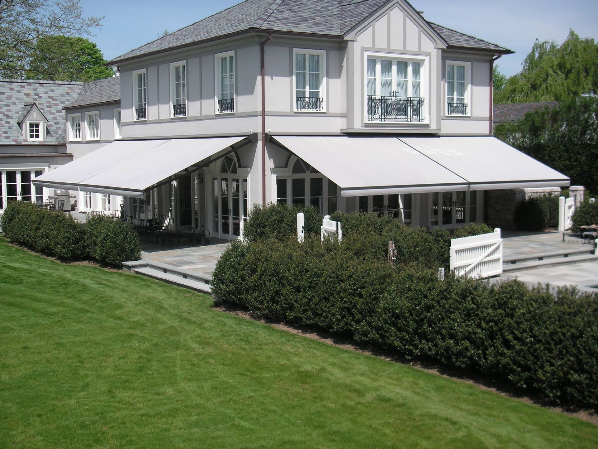 Large house with light-colored retractable awnings over patio, green lawn, and shrubbery.