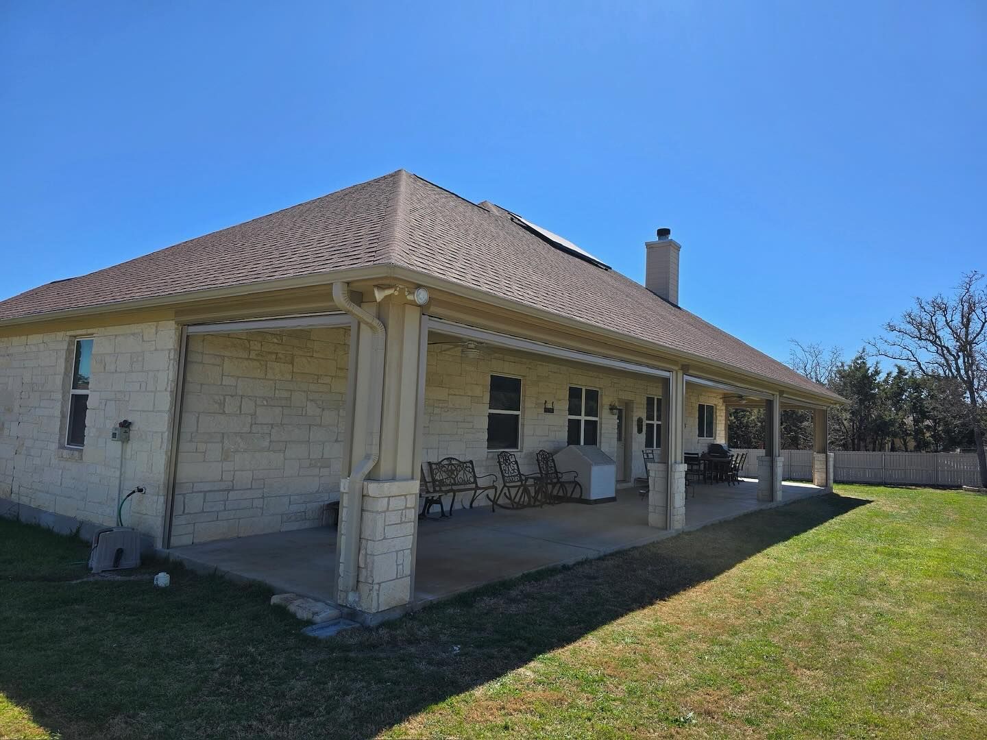 Tan brick house with covered patio, brown roof, and green lawn under a bright blue sky.