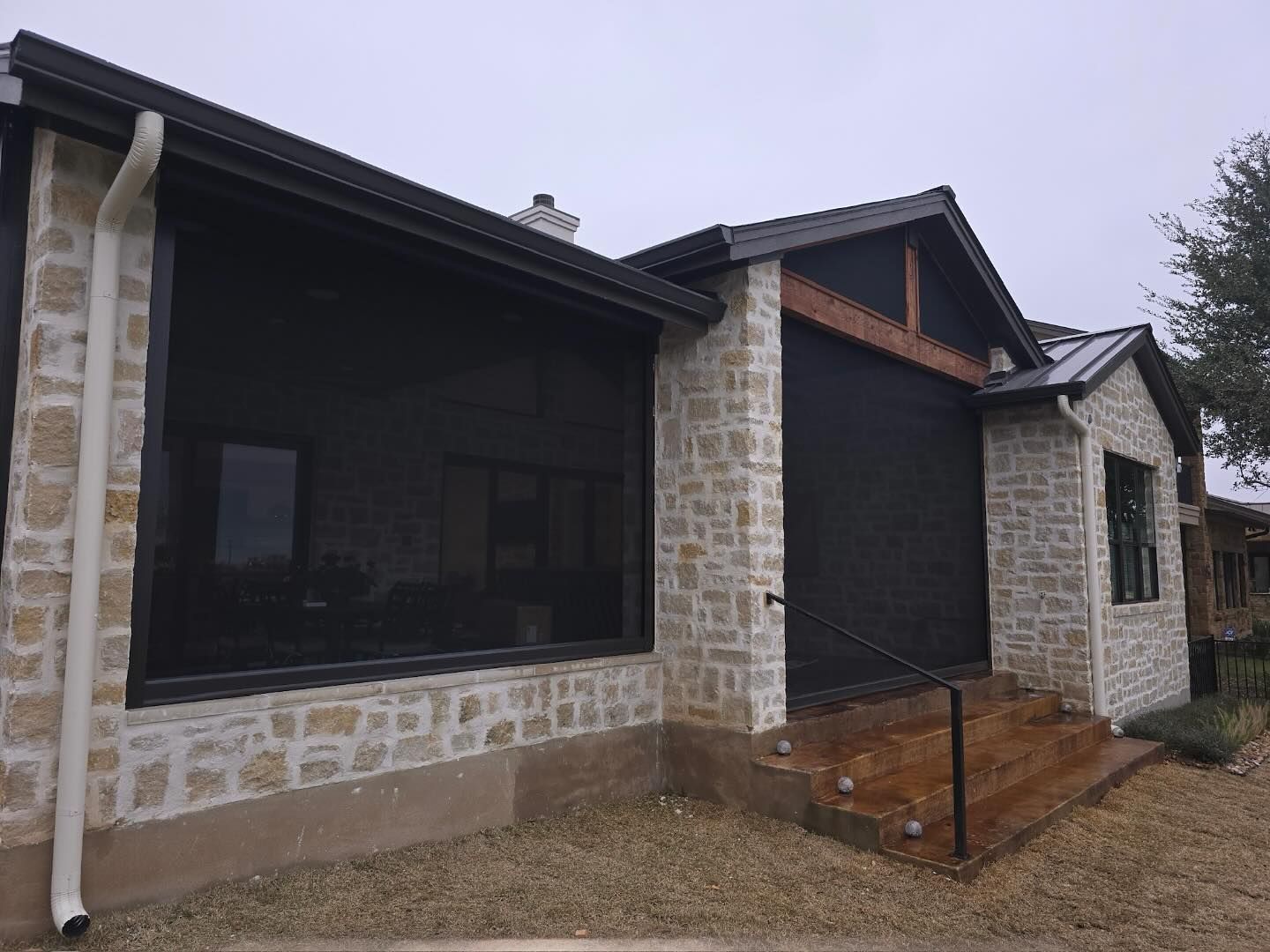 House with stone exterior, black shades covering windows. Rain gutters and gravel ground.