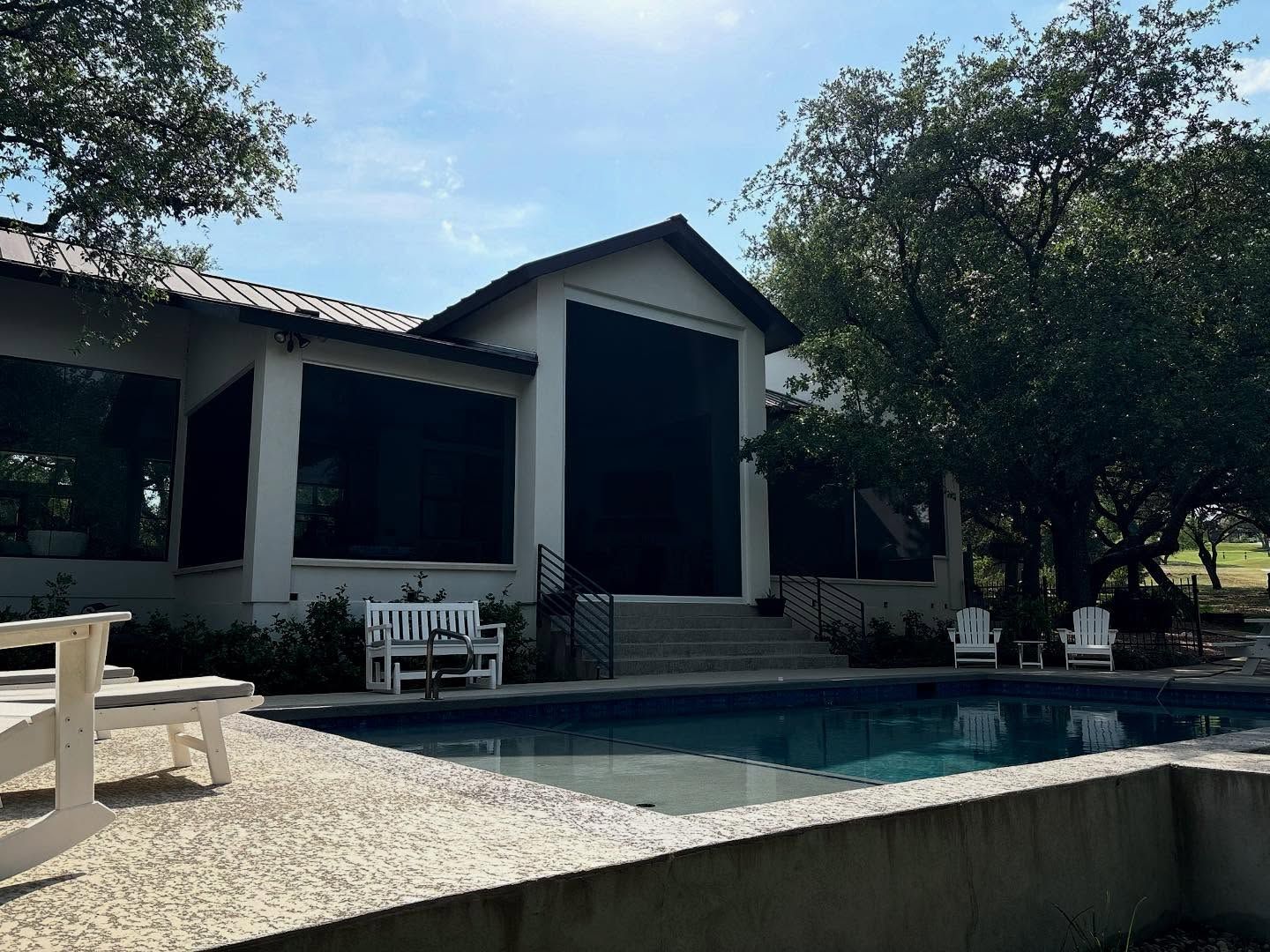 Backyard with pool, patio, and house with black window shades. Trees and sunny sky.
