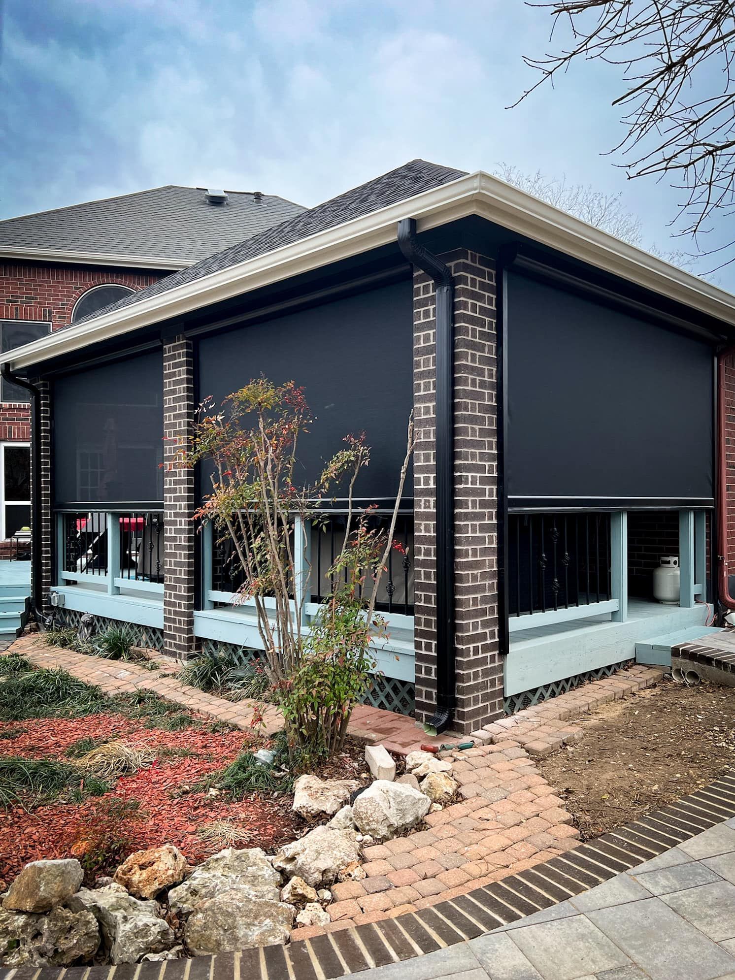 Covered patio with black shades, brick columns, and a landscaped garden.