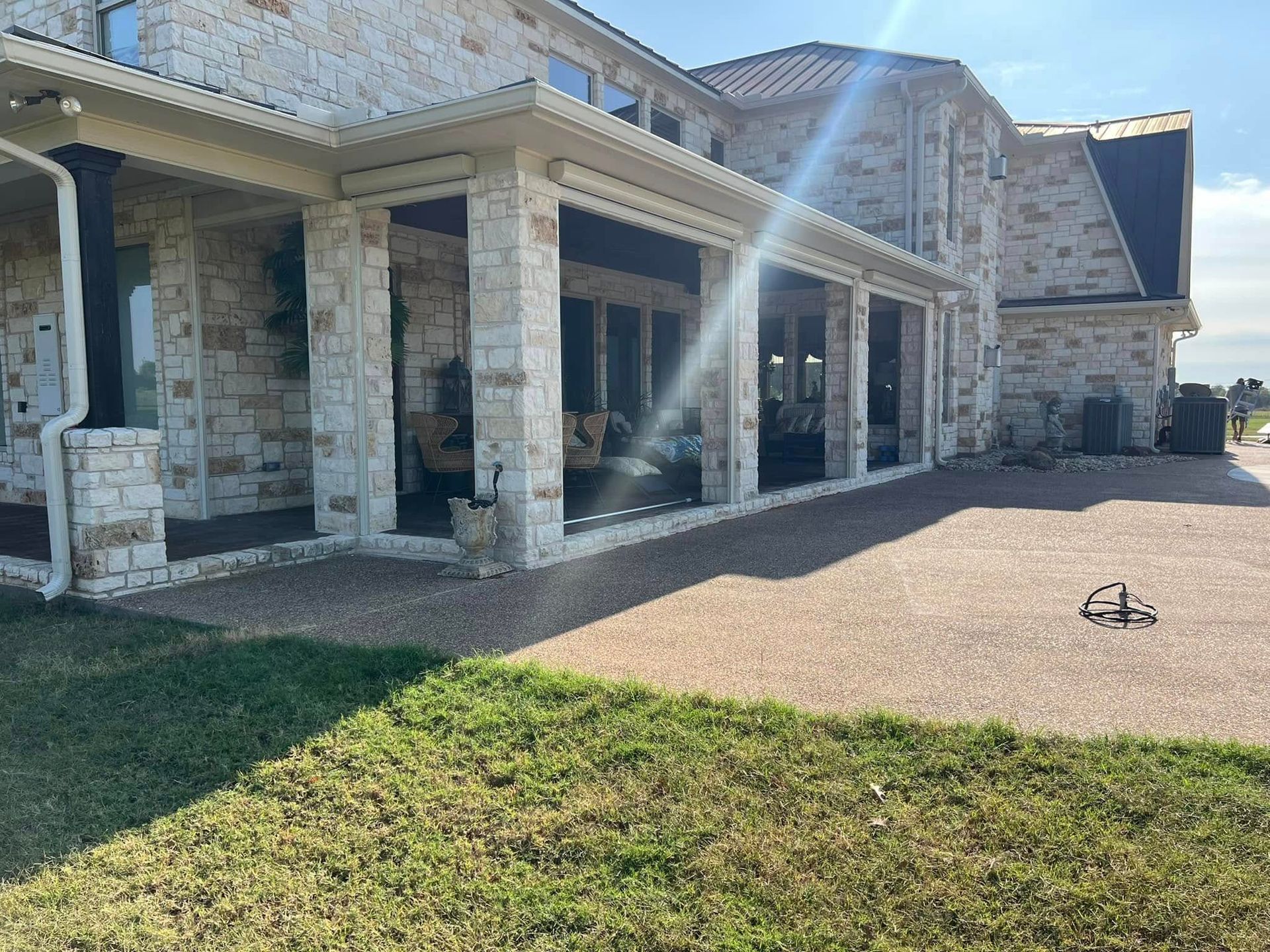 Large stone house with a covered patio, gravel walkway, and grassy yard on a sunny day.