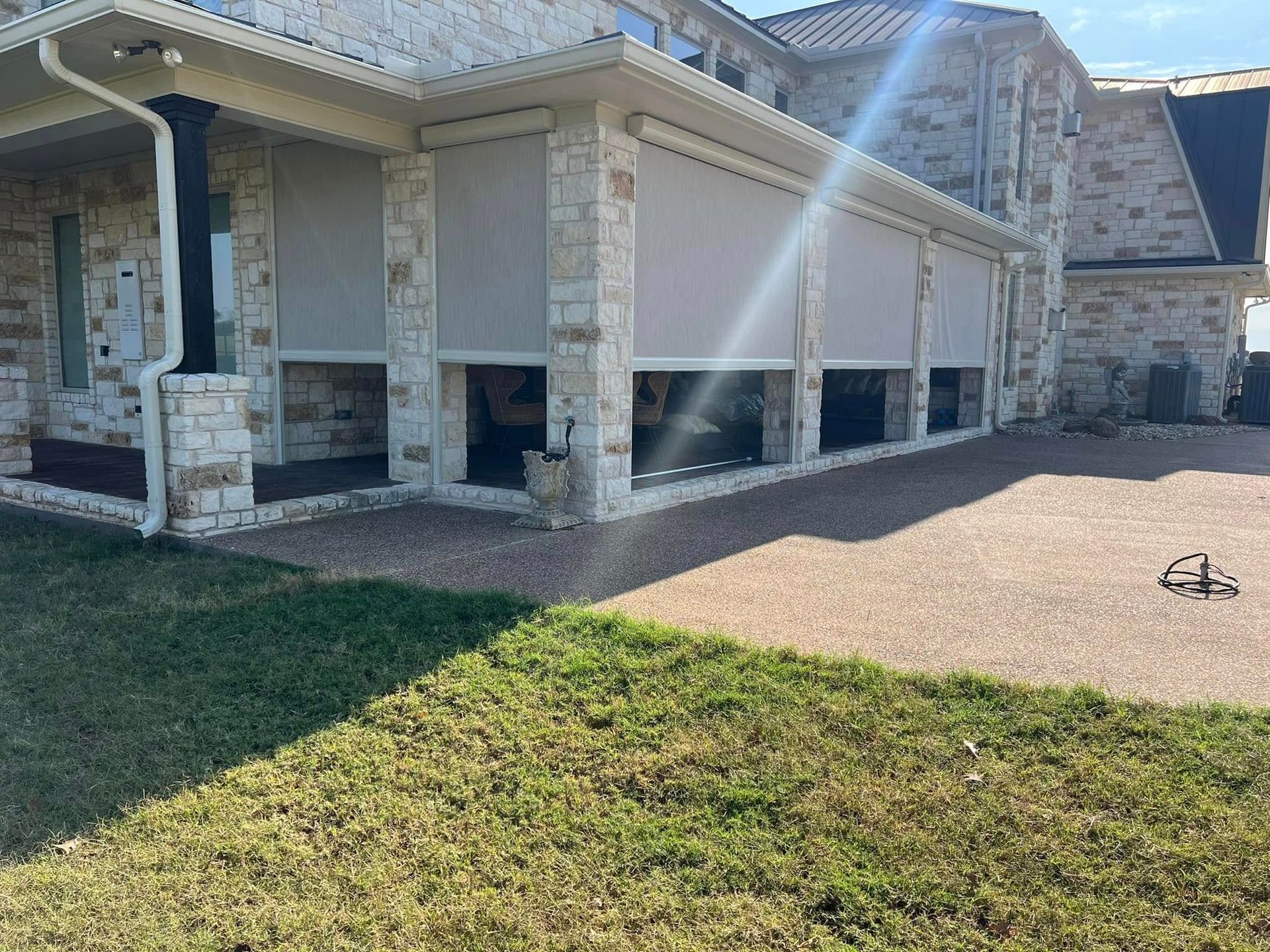 Exterior of a house with stone walls and retractable shades. A concrete patio and green lawn are in the foreground.