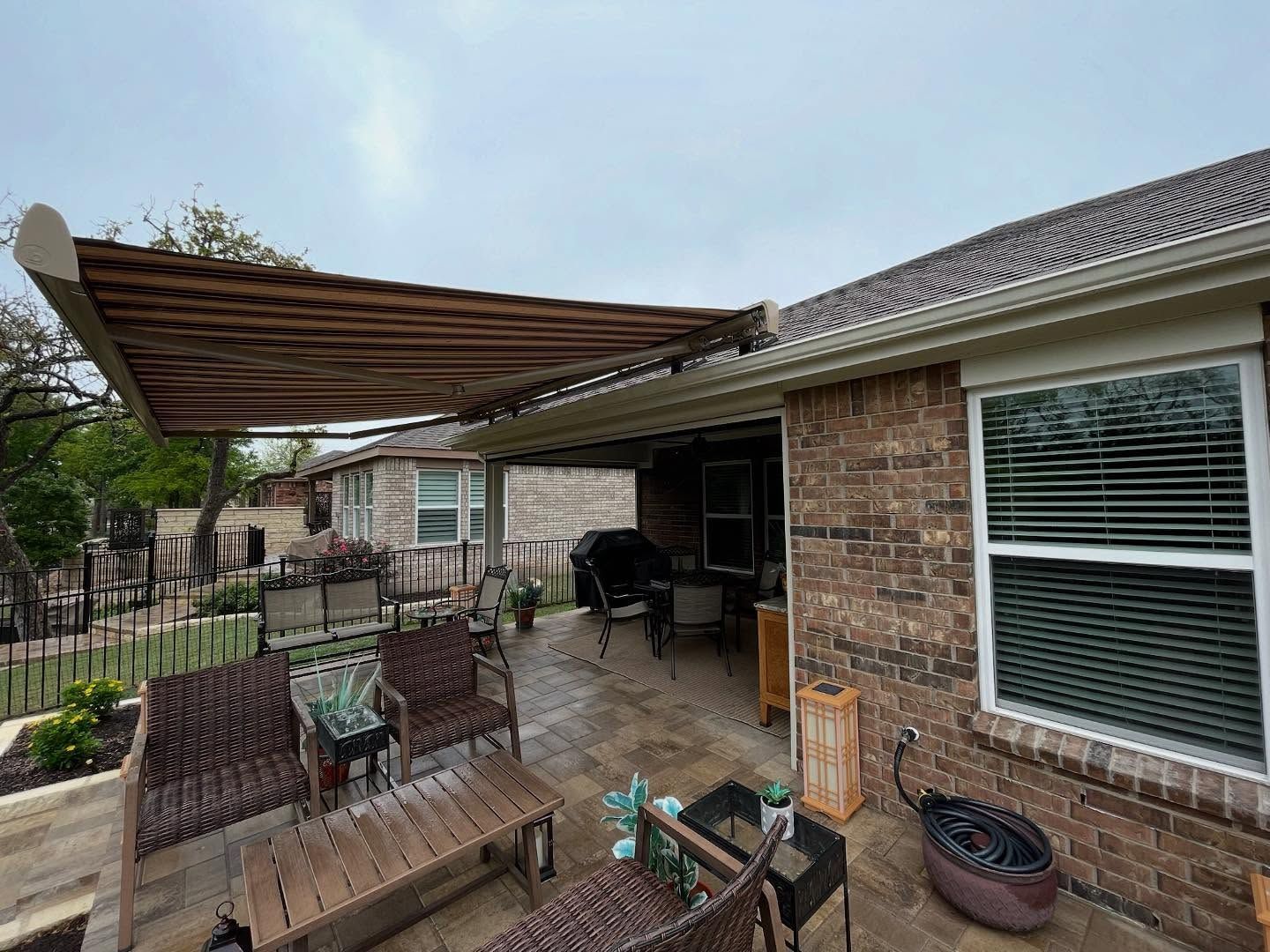 Patio with brown awning, brick house, outdoor furniture, and overcast sky.