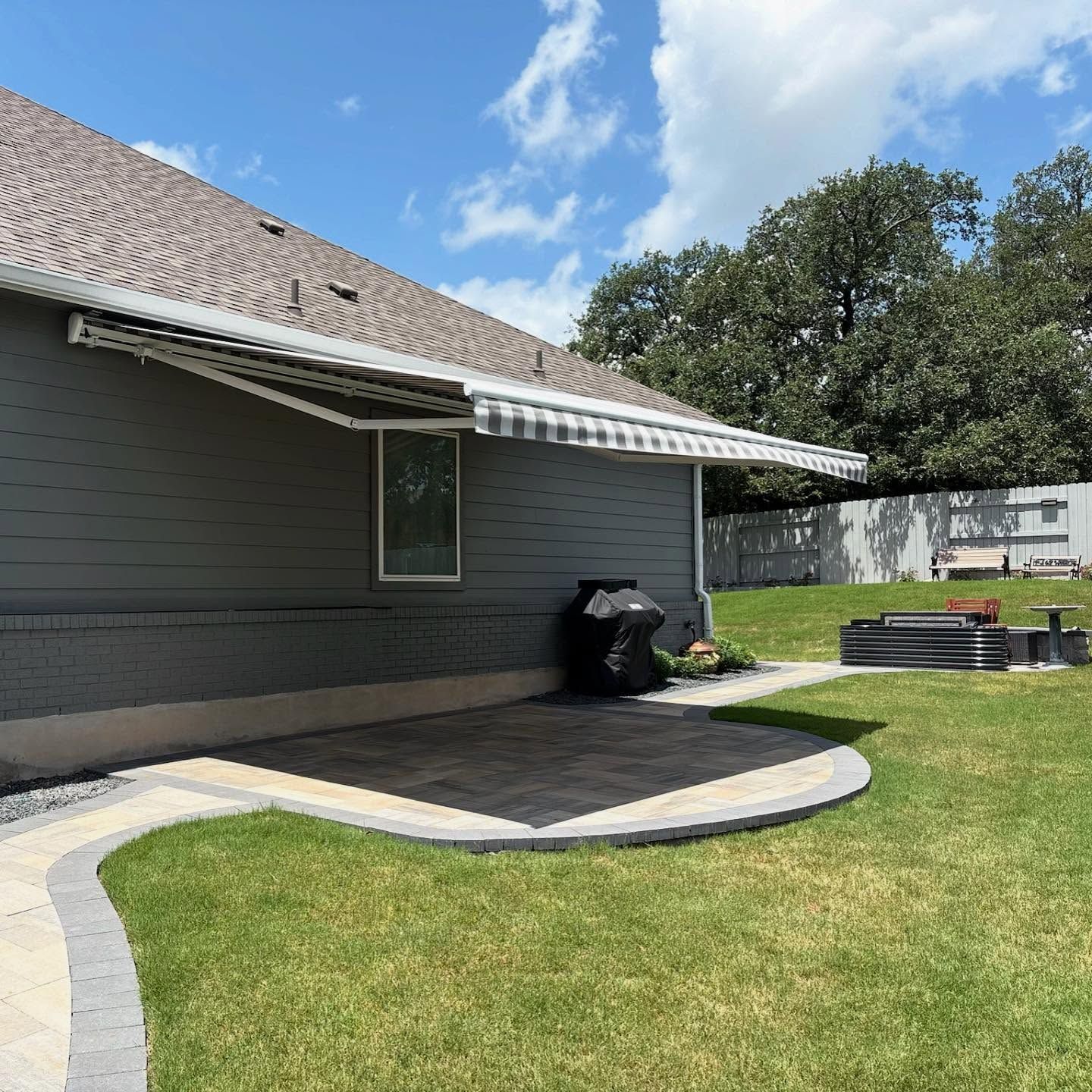 Backyard with an awning over a patio. Gray house with green grass and a path to a fire pit. Sunny day.