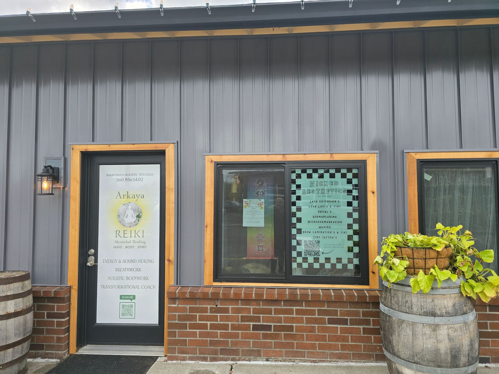 A gray building with a brick wall and barrels in front of it.