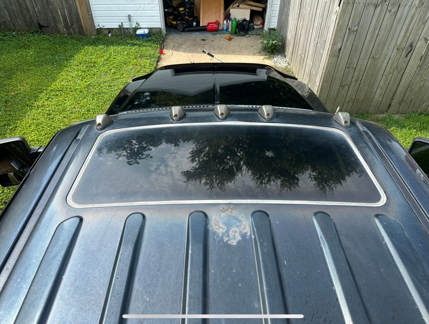 Looking down at a black car roof with sunroof and reflections, parked beside a grassy yard and fence.