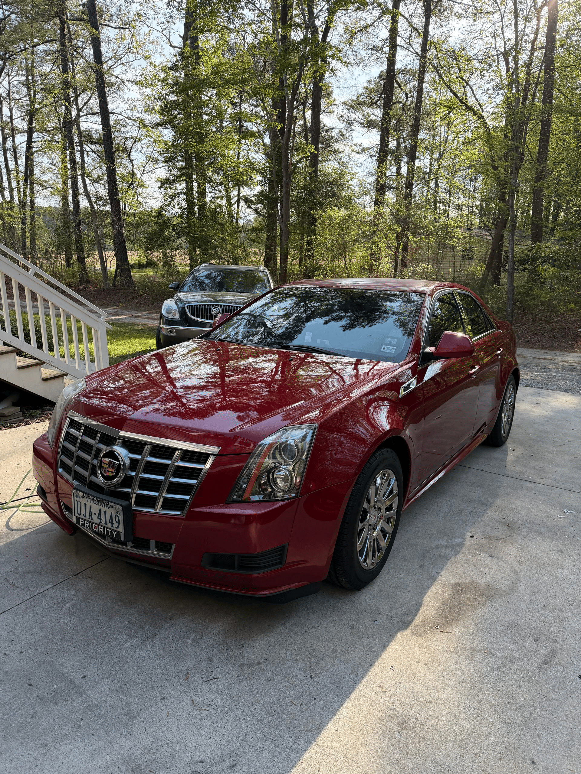 Red Cadillac sedan parked on a driveway beside a wooded yard, with a second car in the background.