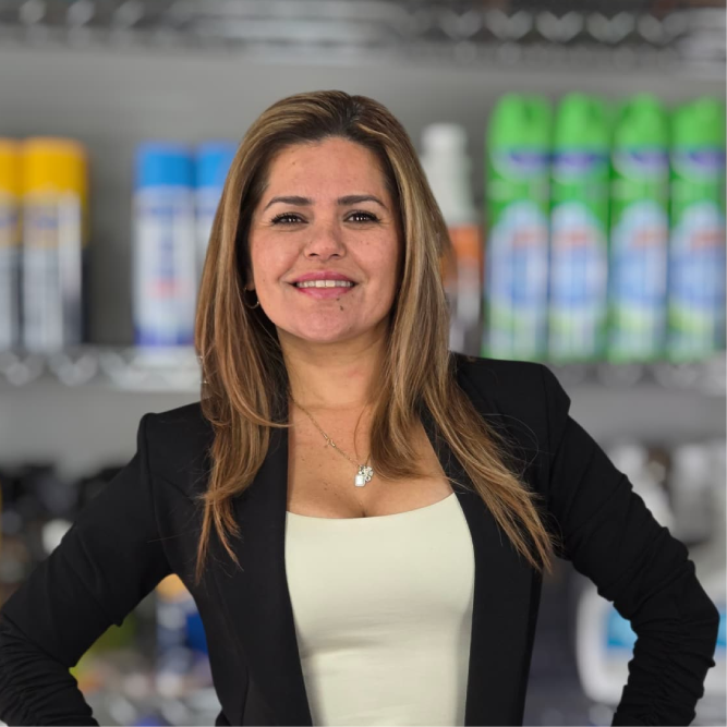 Woman in a blazer smiles, standing in front of shelves stocked with cleaning supplies.
