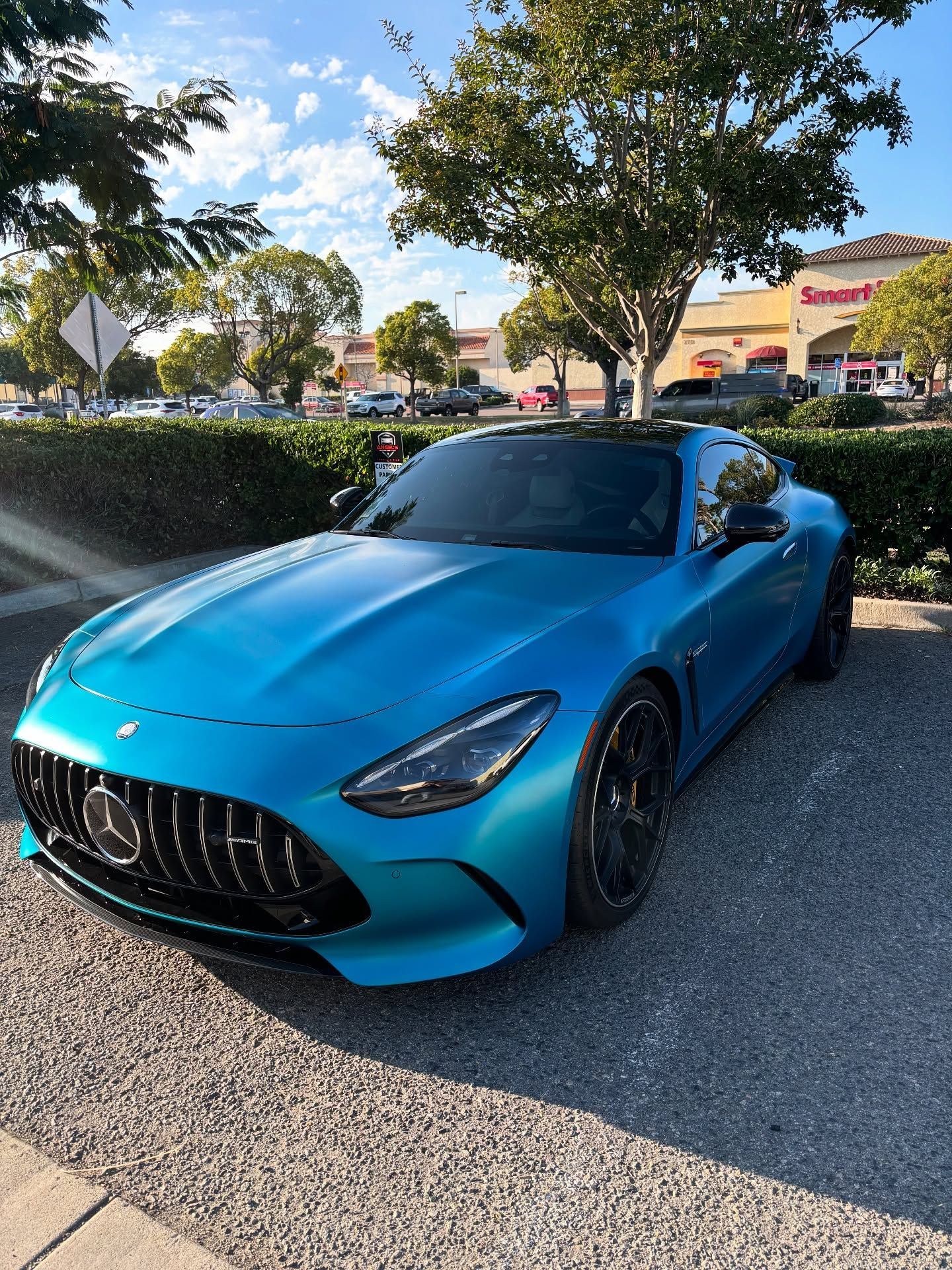Blue Mercedes-AMG GT sports car parked in a parking lot, with a tree and shops visible in the background.