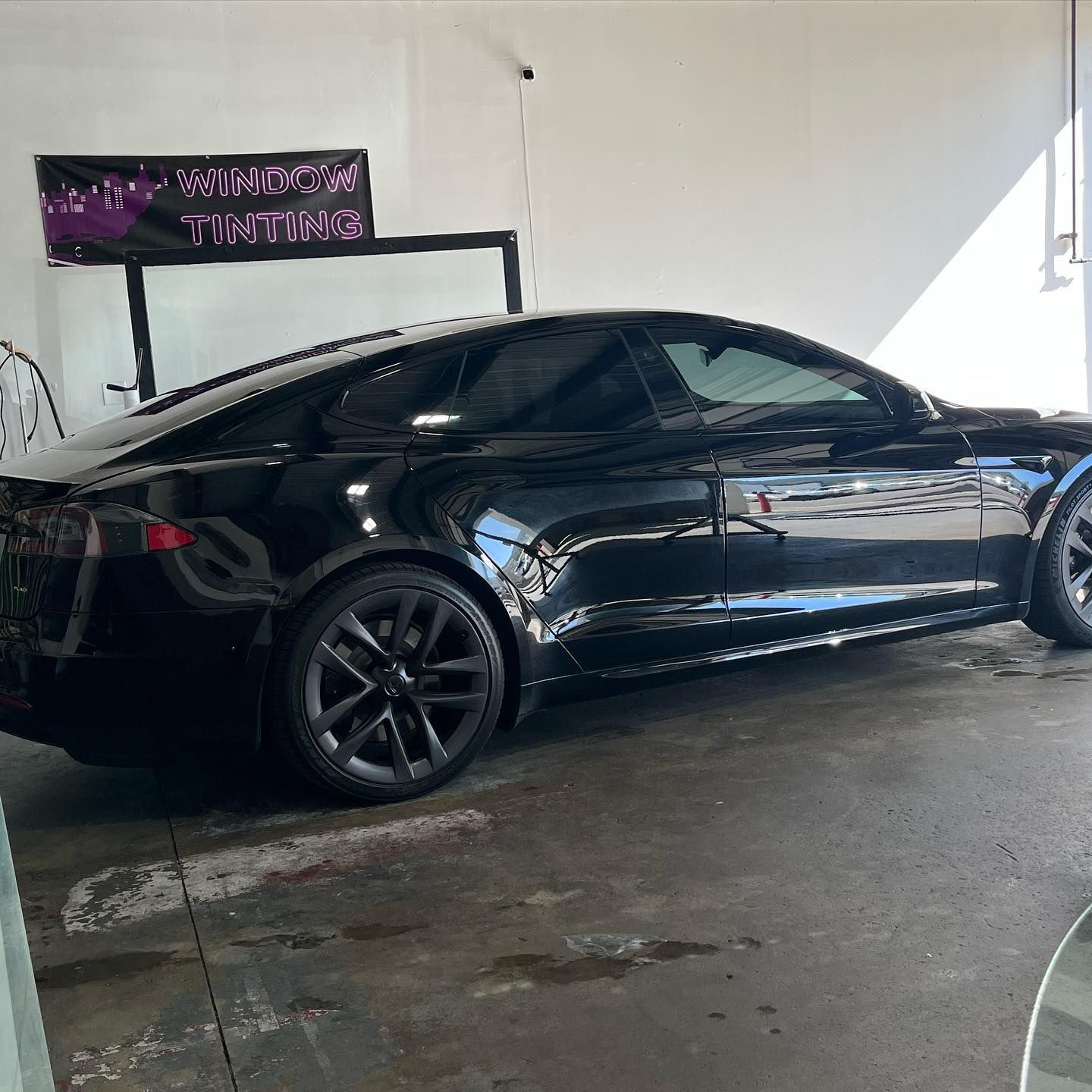 Black Tesla car with tinted windows in a garage, next to a window tinting sign.