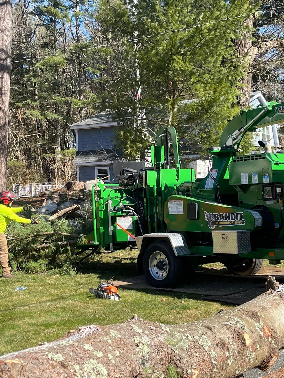 A worker in a bright yellow shirt feeds branches into a large, green wood chipper parked on a lawn near a house.