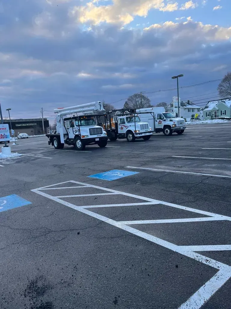 Three white utility bucket trucks parked in a paved, snowy parking lot under a cloudy sky.