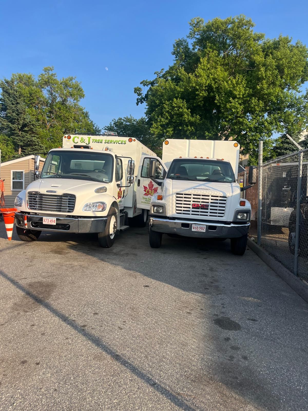 Two white C&J commercial box trucks parked side-by-side on an asphalt lot in front of trees and a chain-link fence.