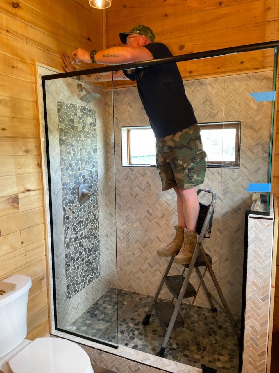 A man is standing on a ladder in a bathroom fixing a shower door.