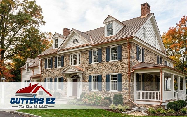 Stone house exterior with blue shutters, autumn trees, and Gutters EZ logo overlay.