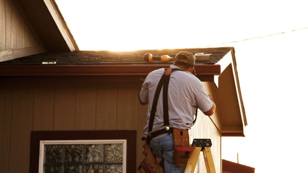 Worker on a ladder installing roof trim on a house during sunset.