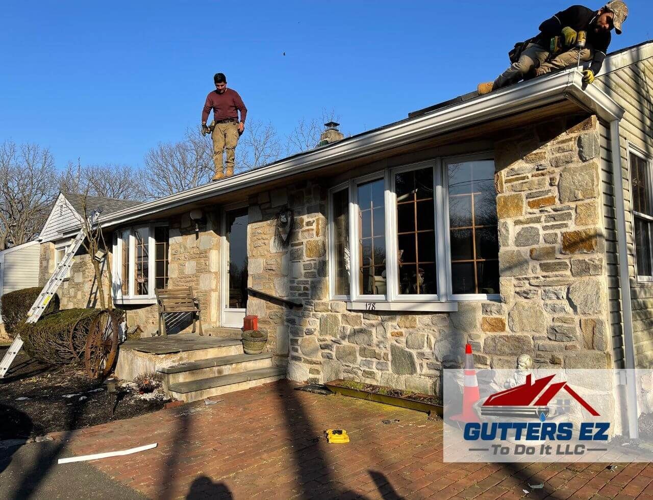 Stone house roof being repaired by workers; gutter company logo visible in lower right.