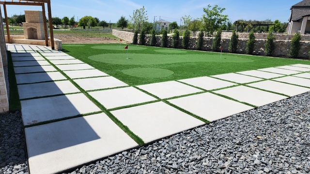 Concrete pavers bordered by turf, leading to a small putting green, with gravel, and trees in the background.