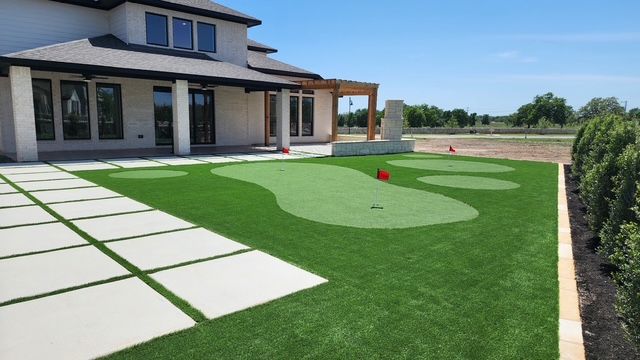 Backyard putting green with a house, sunny day, and concrete patio.