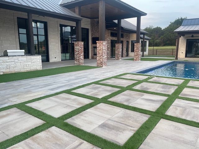 Patio with stone pavers and artificial grass, adjacent to a pool and modern house with brick columns.