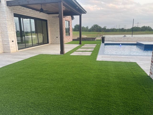 Backyard with green turf, patio, pool, and house with brick accents under a cloudy sky.