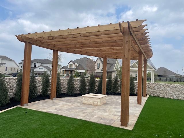 Wooden pergola over a patio with a fire pit, surrounded by green lawn, shrubs, and stone walls.