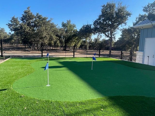 Green putting green with three blue flags, blue-sided building in the background, trees, and clear sky.