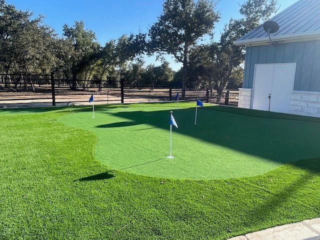 A backyard putting green with three flags. Bright green turf, blue flags, near a light blue building.