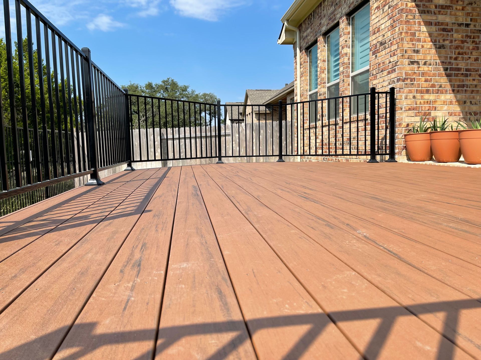 A wooden deck with a metal railing and potted plants in front of a brick house.