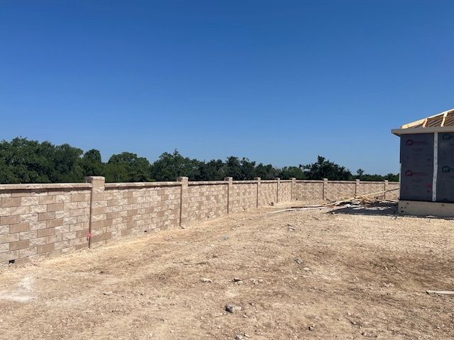 A brick wall surrounds a dirt field with a house in the background.