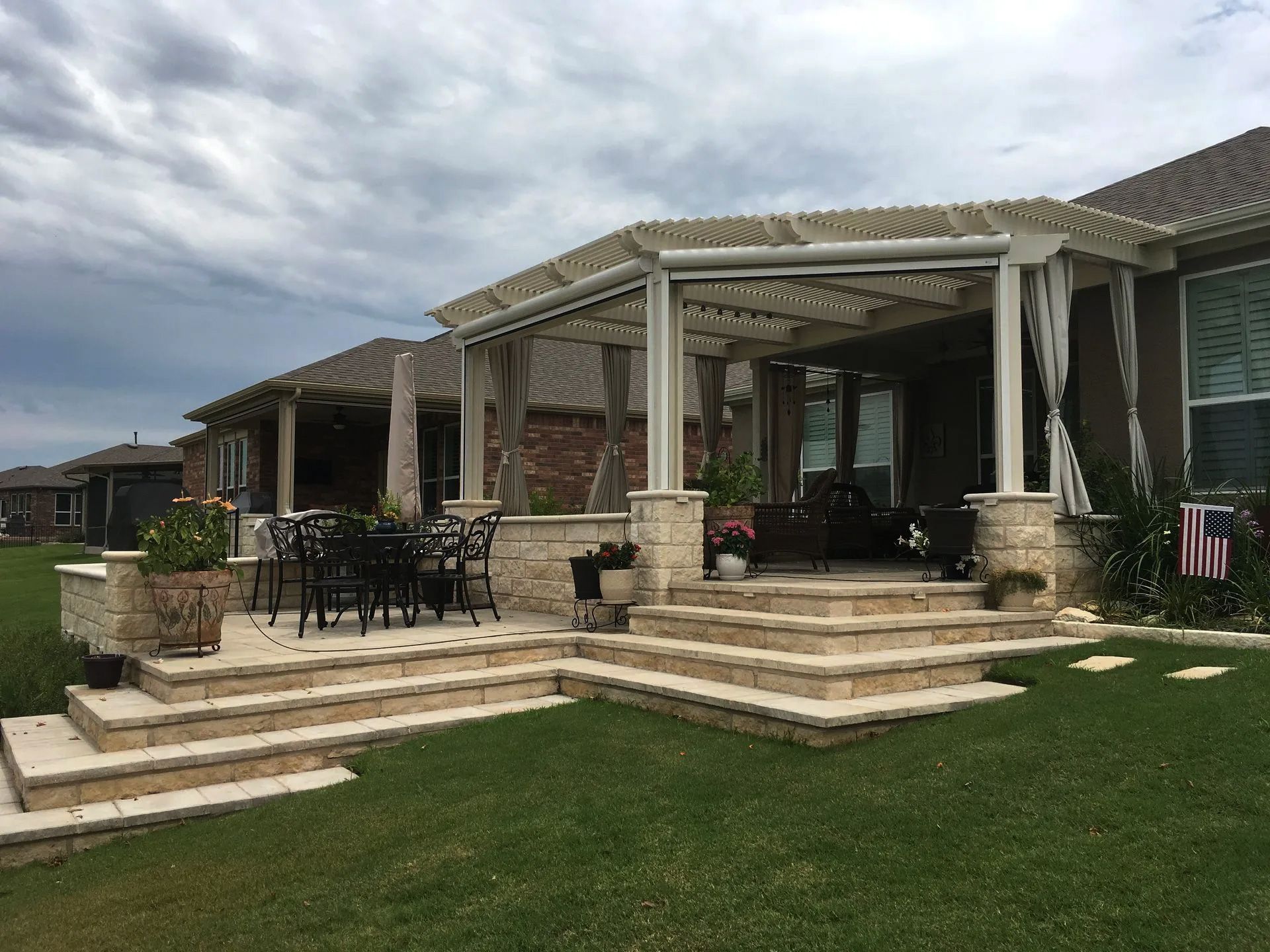 Stone patio with pergola, steps, and outdoor seating in a backyard. Green grass and cloudy sky.